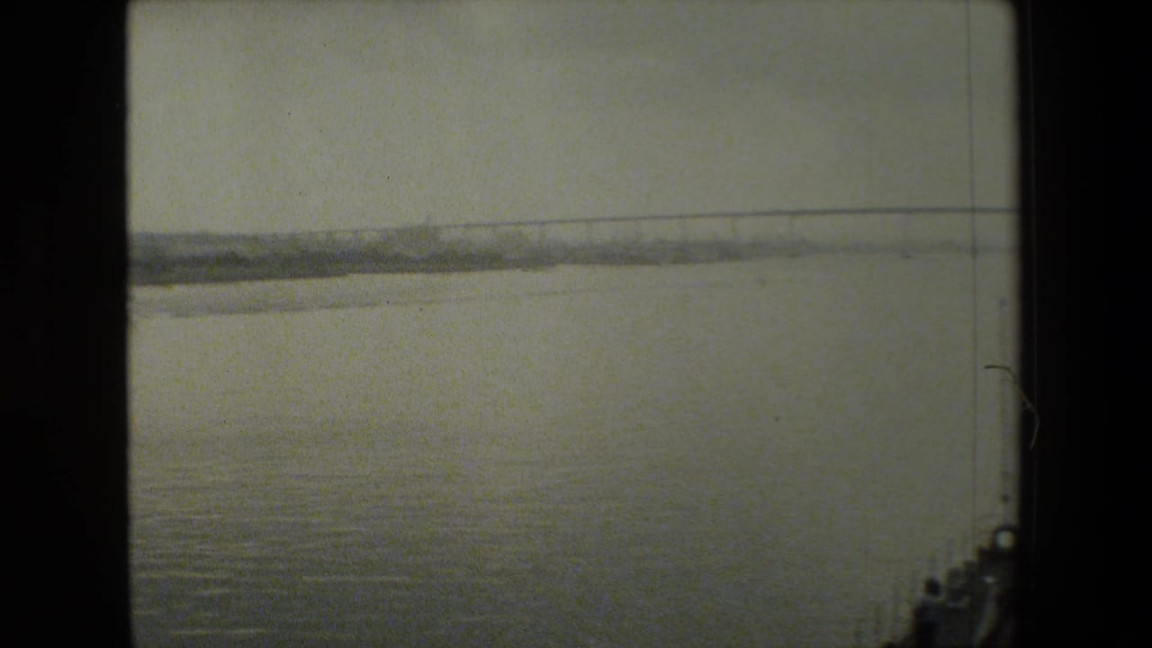 A hazy 8mm view from a moving boat shows a broad water surface, a long bridge in the distance, and part of the vessel at the right frame edge.