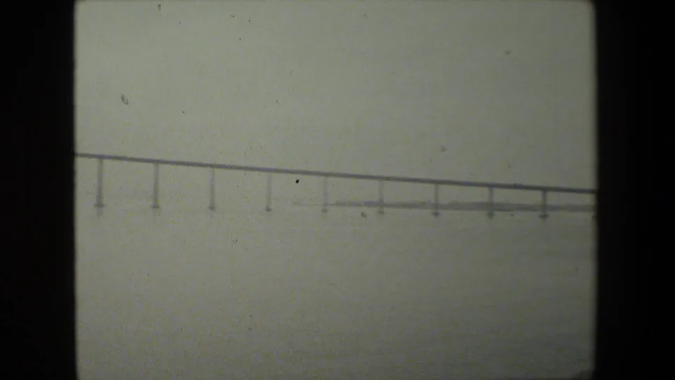 A soft 8mm frame shows a long low bridge stretching across a hazy waterfront, with open water below and dark film borders at the sides.