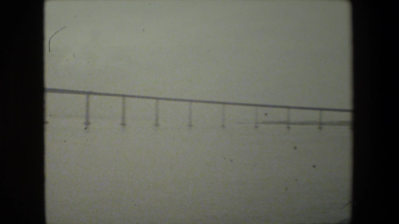 Soft 8mm frame showing a distant low bridge or elevated span over water, with repeated vertical supports and a hazy overcast background.