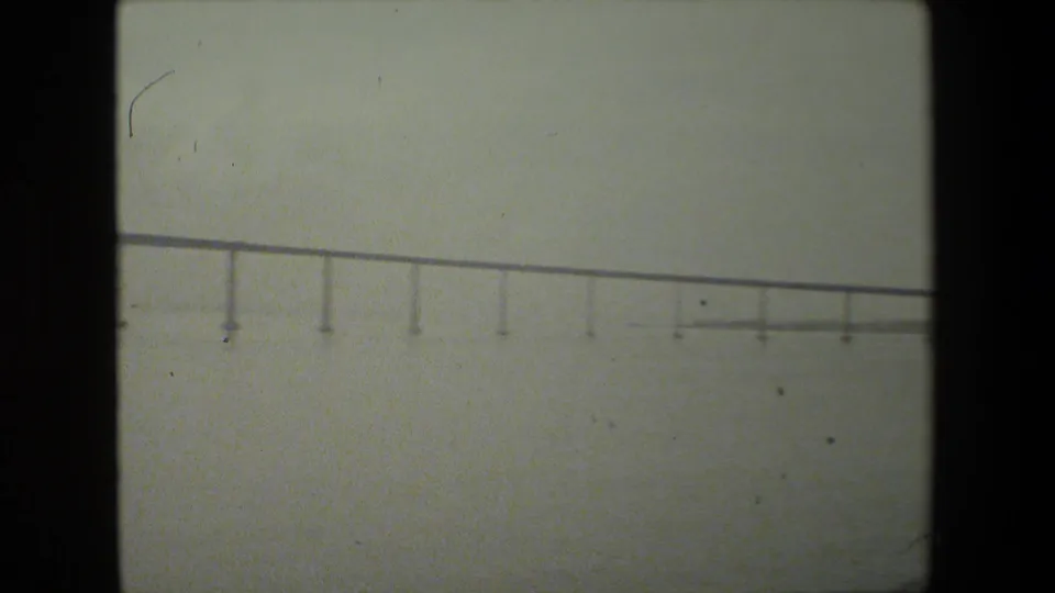Soft 8mm frame showing a distant low bridge or elevated span over water, with repeated vertical supports and a hazy overcast background.