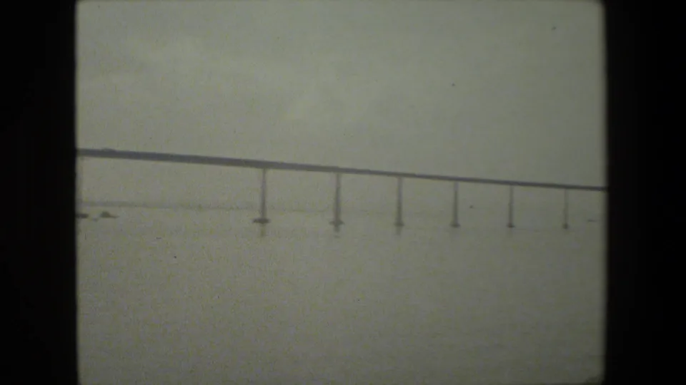 A long horizontal bridge structure appears across open water, with small pier supports and soft grain from the 8mm scan.