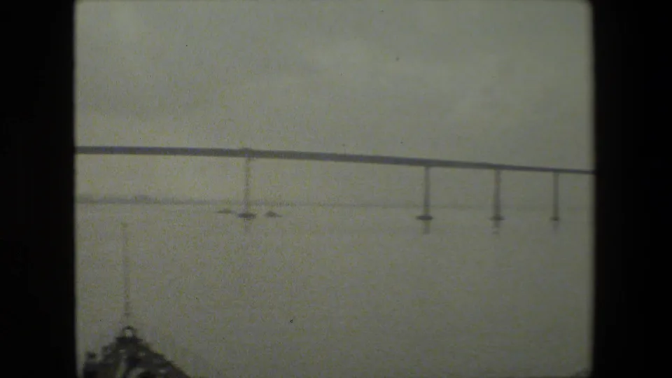 The camera view moves closer to a low bridge span, showing vertical supports descending toward calm water and a faint shoreline in the distance.