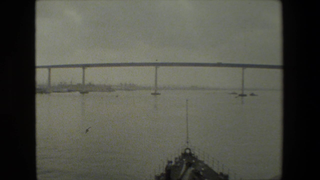 A boat-level view shows the vessel bow in the foreground with a long bridge spanning open water and a hazy shoreline beyond, from archival 8mm footage described as near a naval base.