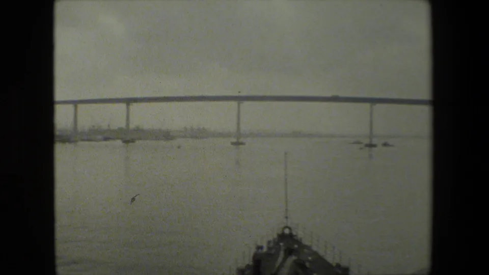 A boat-level view shows the vessel bow in the foreground with a long bridge spanning open water and a hazy shoreline beyond, from archival 8mm footage described as near a naval base.