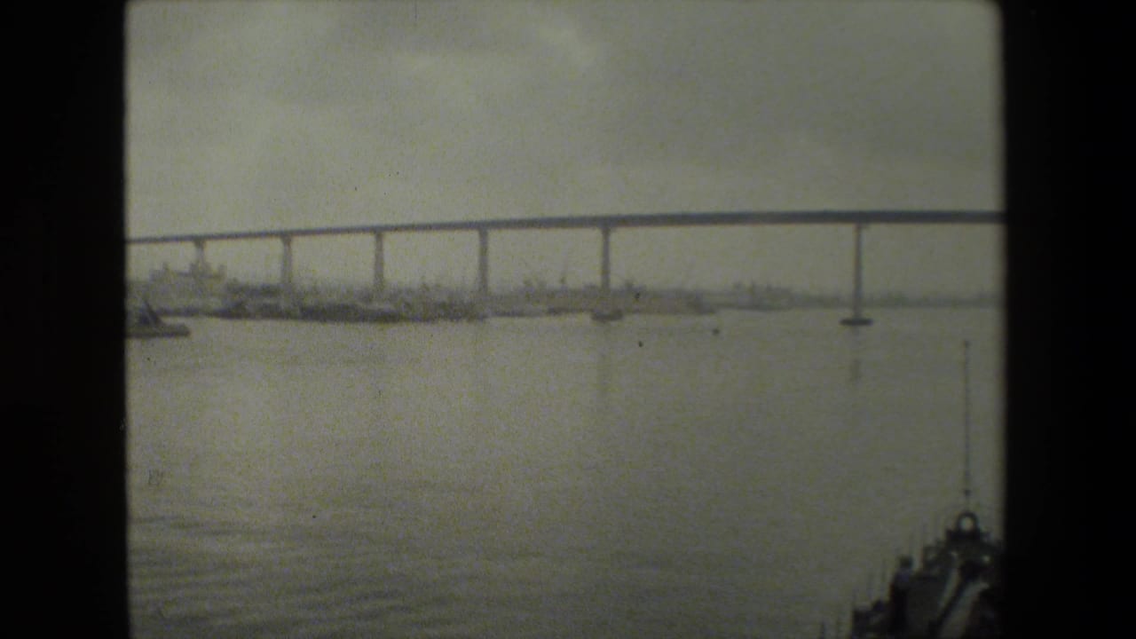 The boat bow remains at the lower right while a long bridge crosses the background, with small boats or buoys visible on the water beneath a muted sky.