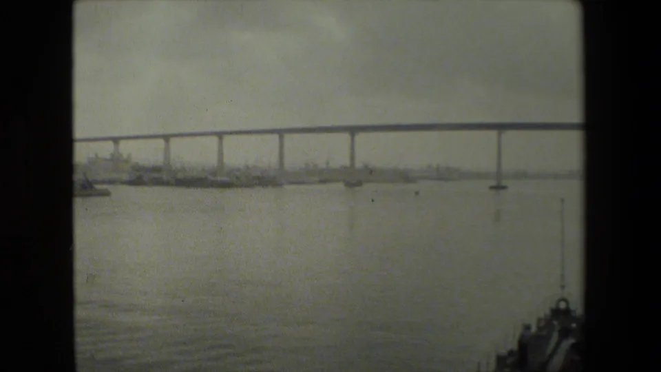 The boat bow remains at the lower right while a long bridge crosses the background, with small boats or buoys visible on the water beneath a muted sky.