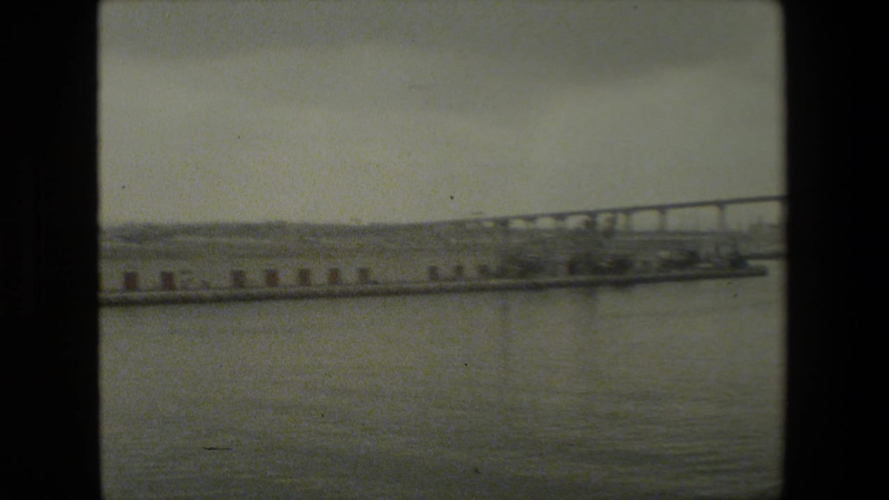 Soft 8mm frame of a waterfront scene with open water in the foreground, a long pier across the middle distance, and a bridge or causeway receding along the horizon.