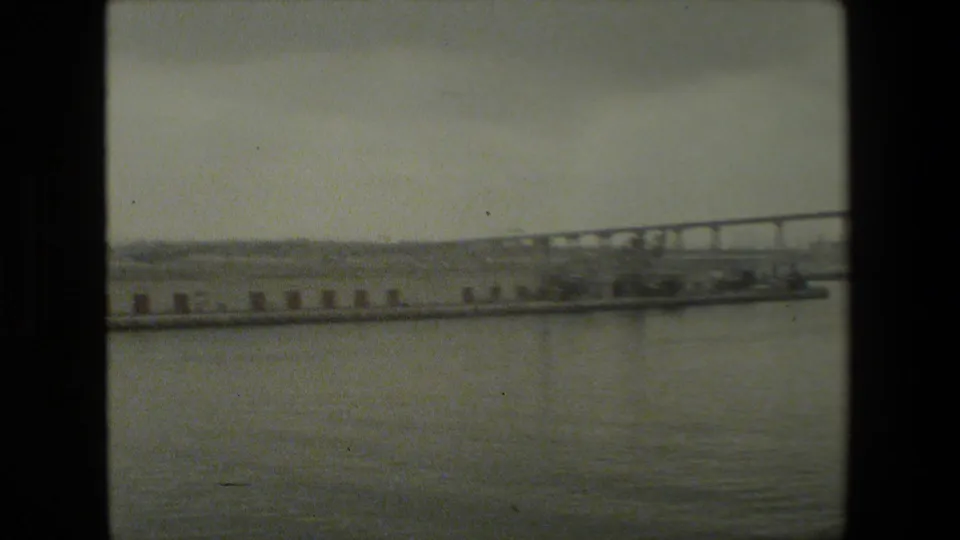 Soft 8mm frame of a waterfront scene with open water in the foreground, a long pier across the middle distance, and a bridge or causeway receding along the horizon.