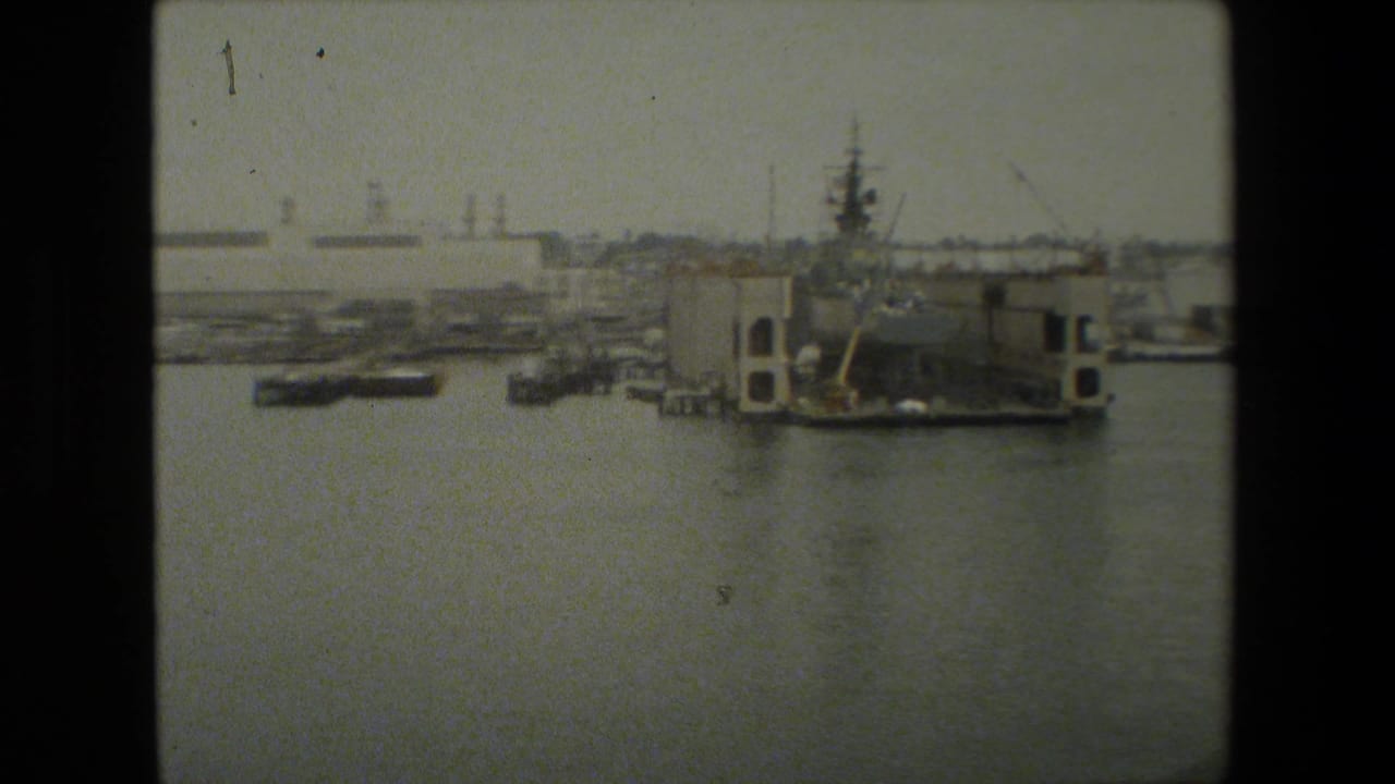 Archival 8mm view across harbor water toward small boats, dockside structures, cranes, and a large ship or drydock-side structure near the right edge of the frame.