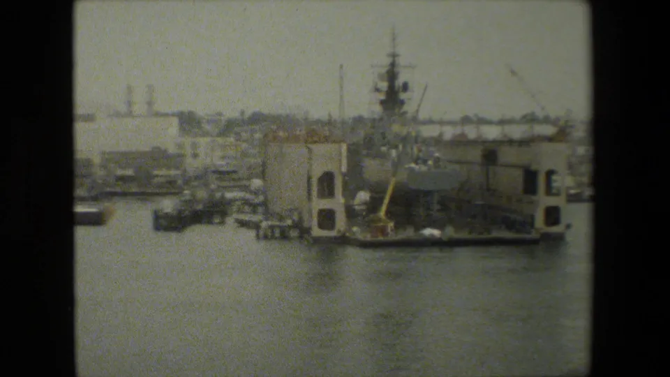 Soft, grainy 8mm frame looking across water toward moored boats, cranes, dock buildings, and a large vessel or maintenance structure along the waterfront.
