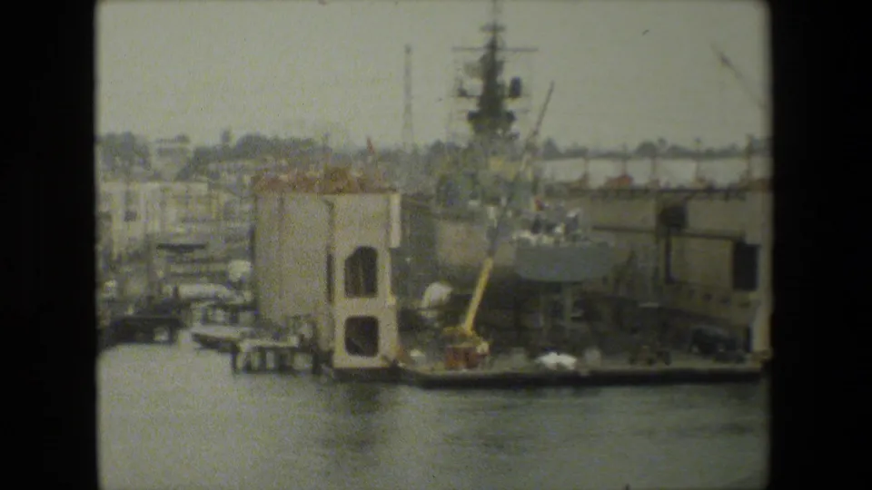 Archival 8mm frame of a waterfront maintenance area with a large ship or drydock-side structure, cranes, dock buildings, small boats, and harbor water in the foreground.