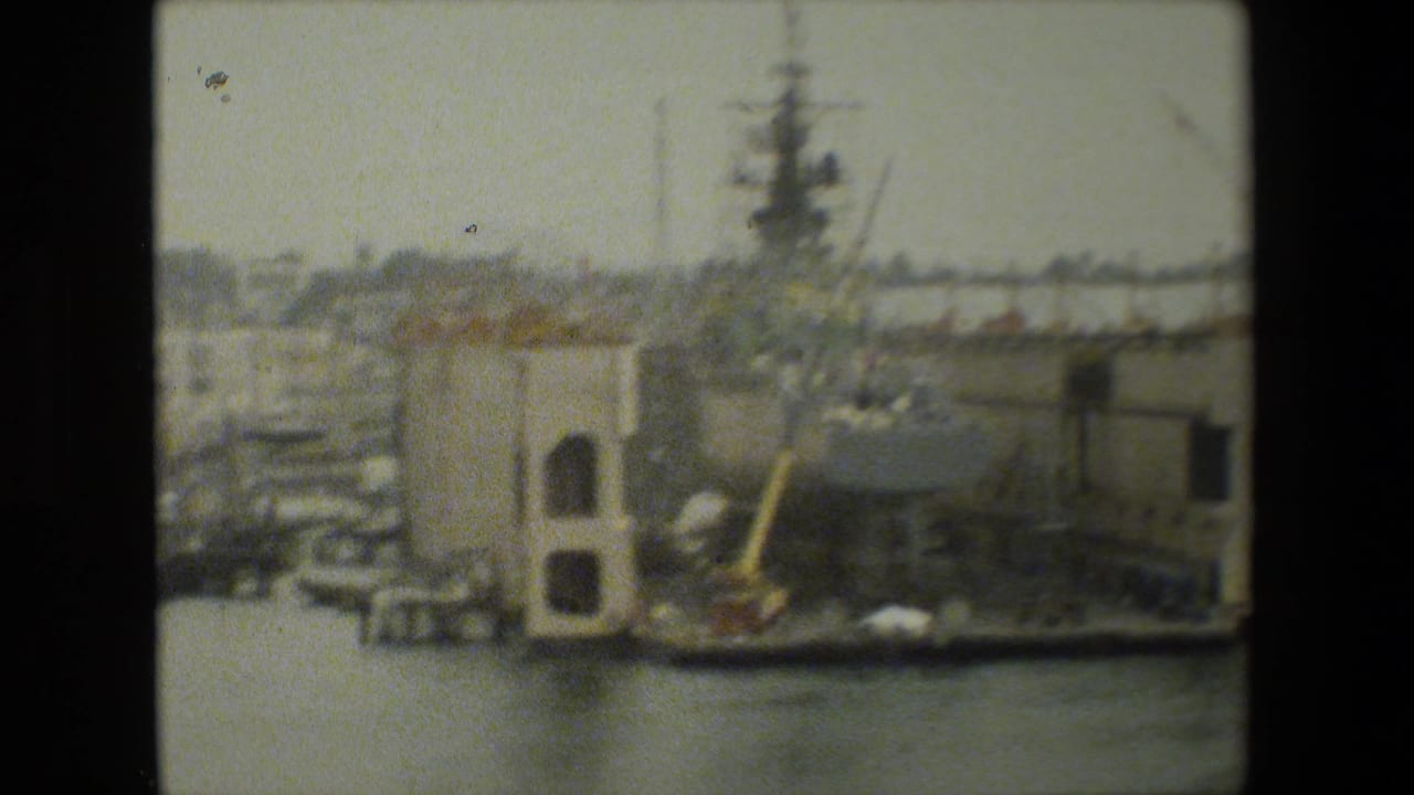 Soft 8mm frame of a large dockside vessel or industrial pier structure beside calm harbor water, with cranes and naval infrastructure faintly visible behind it.