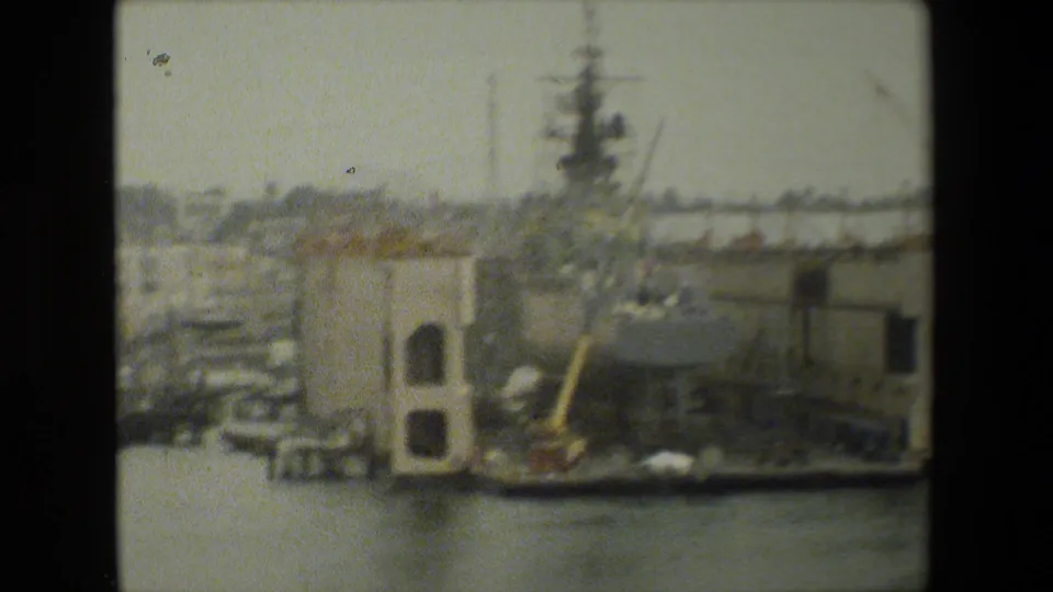 Soft 8mm frame of a large dockside vessel or industrial pier structure beside calm harbor water, with cranes and naval infrastructure faintly visible behind it.
