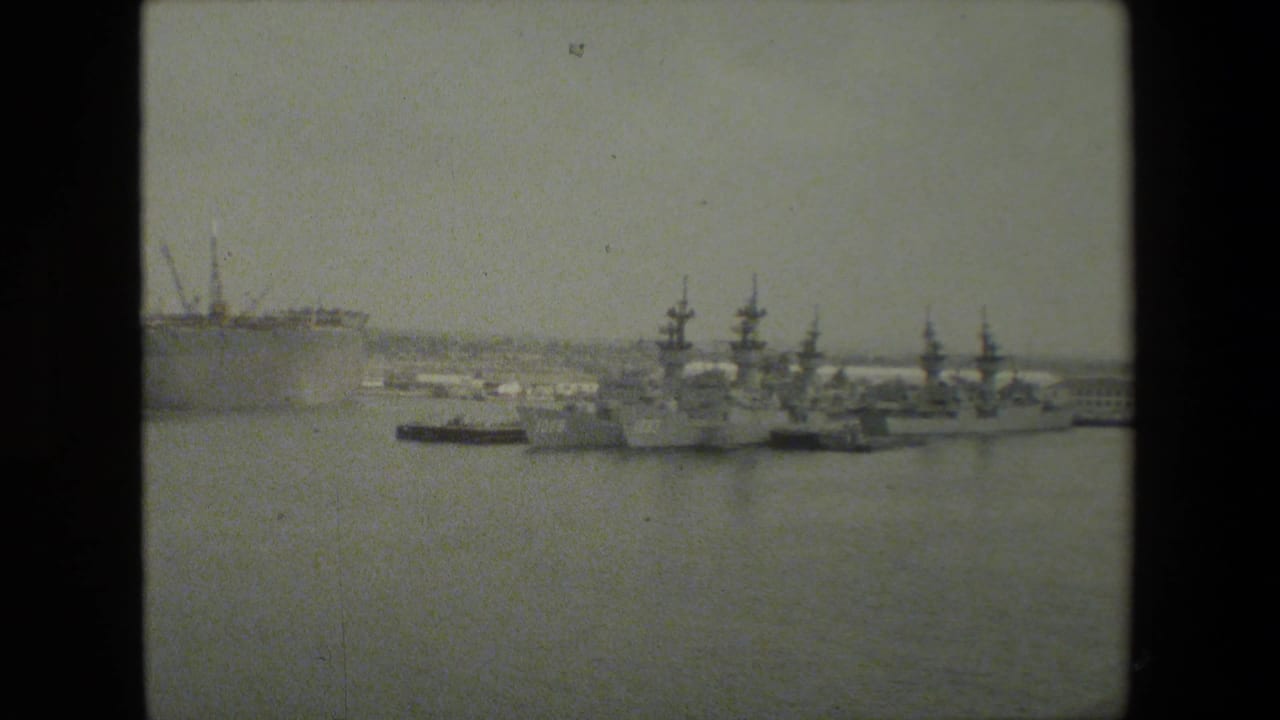 Distant naval ships lined up at a pier across calm harbor water.