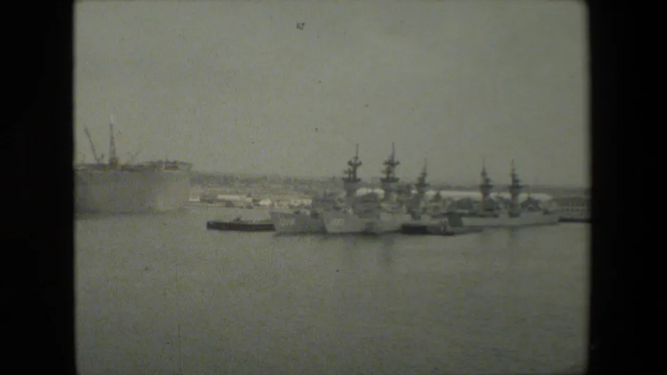 Distant view of multiple gray naval vessels moored along a pier, with tall masts and low harbor buildings under a hazy sky.
