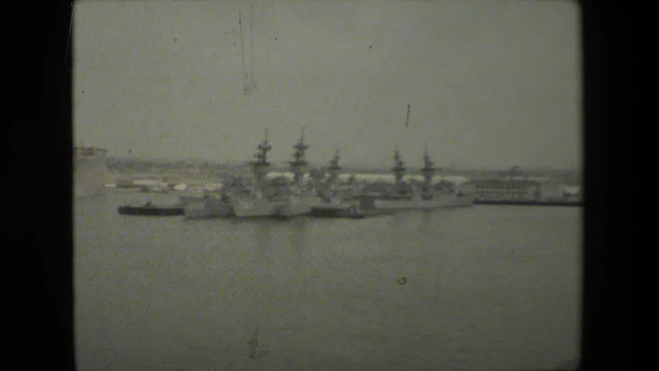 Wide 8mm harbor view showing several docked naval vessels, pier structures, cranes, and waterfront buildings across still water.