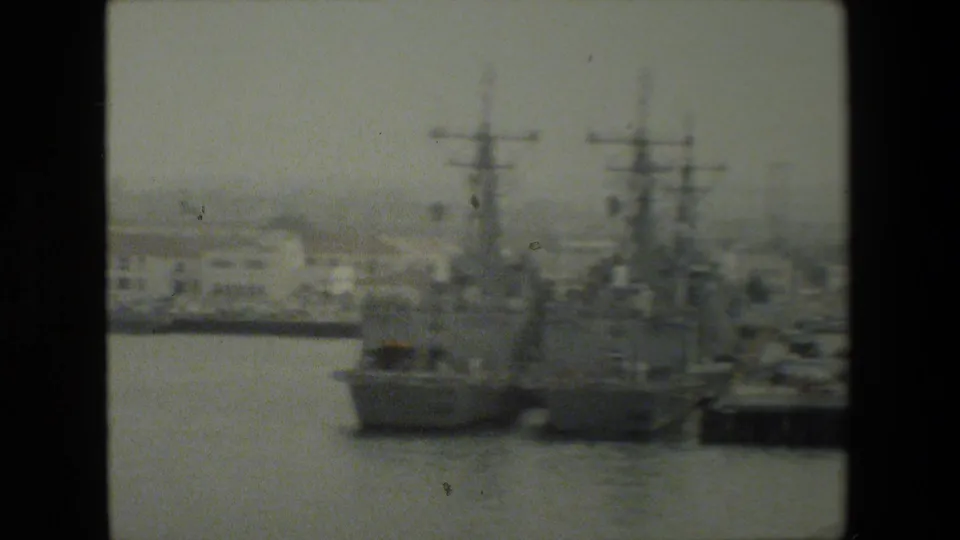 Soft-focus 8mm frame of naval ships moored near a pier, with masts, rigging, and a low waterfront building visible through haze and film grain.