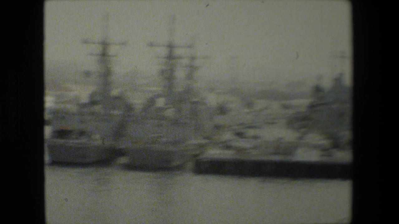 Soft 8mm frame showing multiple docked U.S. Navy-type warships across harbor water, with tall masts, piers, and distant waterfront structures visible.