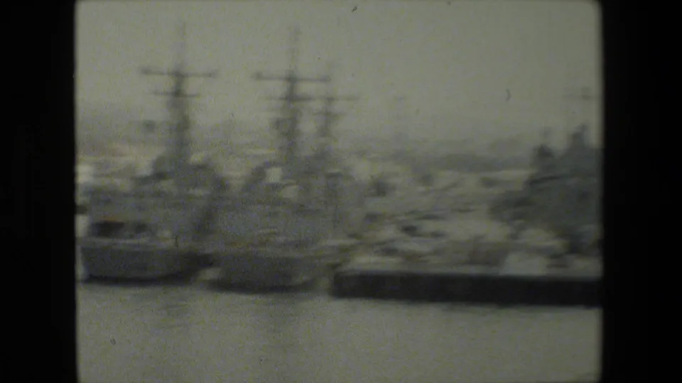 Soft 8mm frame showing multiple docked U.S. Navy-type warships across harbor water, with tall masts, piers, and distant waterfront structures visible.