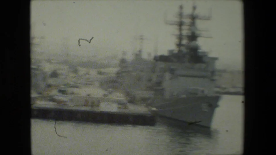 A large naval vessel occupies the right side of the frame while additional moored ships, piers, and harbor water extend into the background.