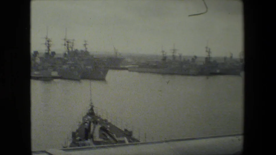 Wide home-movie view of docked naval vessels across calm harbor water, with a foreground roofline or dock structure at the bottom of the frame.