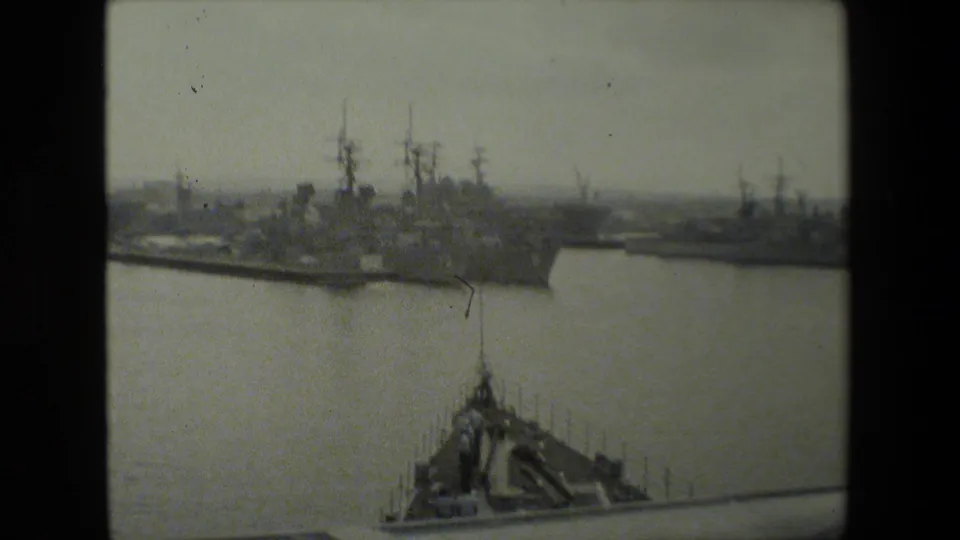 Clustered naval ships and mast silhouettes sit across the harbor, with a lower foreground roofline or dock structure partially framing the water.