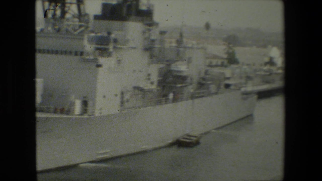 Large gray naval vessel alongside a pier at a harbor, with other ship structures faintly visible in the background in archival 8mm footage.