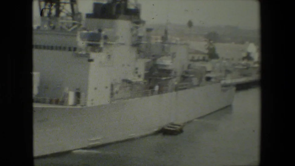 Large gray naval vessel alongside a pier at a harbor, with other ship structures faintly visible in the background in archival 8mm footage.