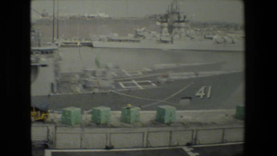 Gray military vessel with a marked deck and the number 41 visible beside green dockside containers and other ships in the harbor.