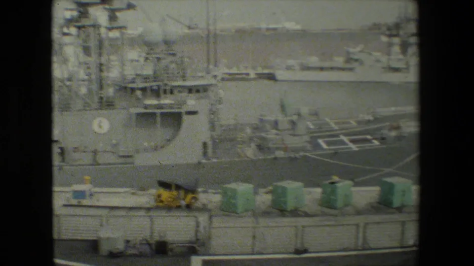 Naval harbor view with a gray military ship, marked deck area, green containers on the pier, and small figures near the dockside equipment.