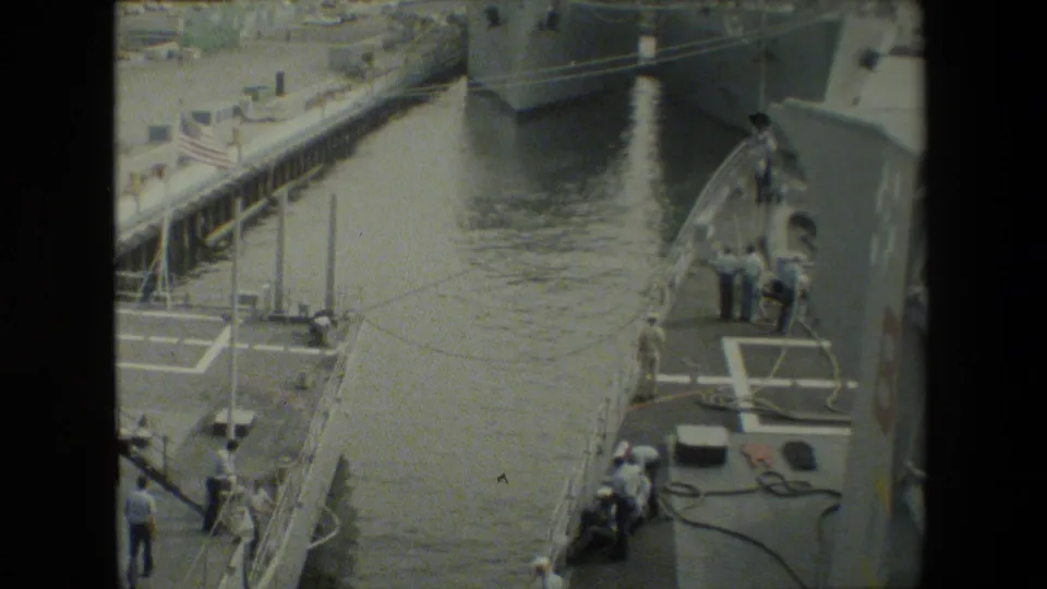 Small crew members or visitors stand near a gangway and deck markings between docked gray naval vessels, with harbor water and pier structures visible.