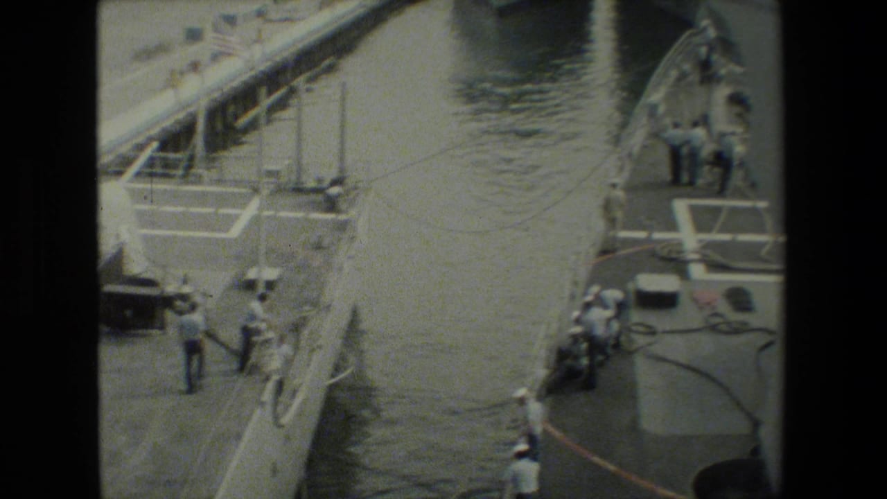 High-angle 8mm frame of a gray military ship moored beside dock water, with sailors in white uniforms working near lines and deck equipment.
