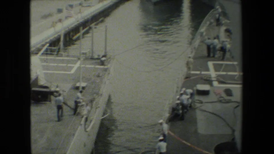 High-angle 8mm frame of a gray military ship moored beside dock water, with sailors in white uniforms working near lines and deck equipment.