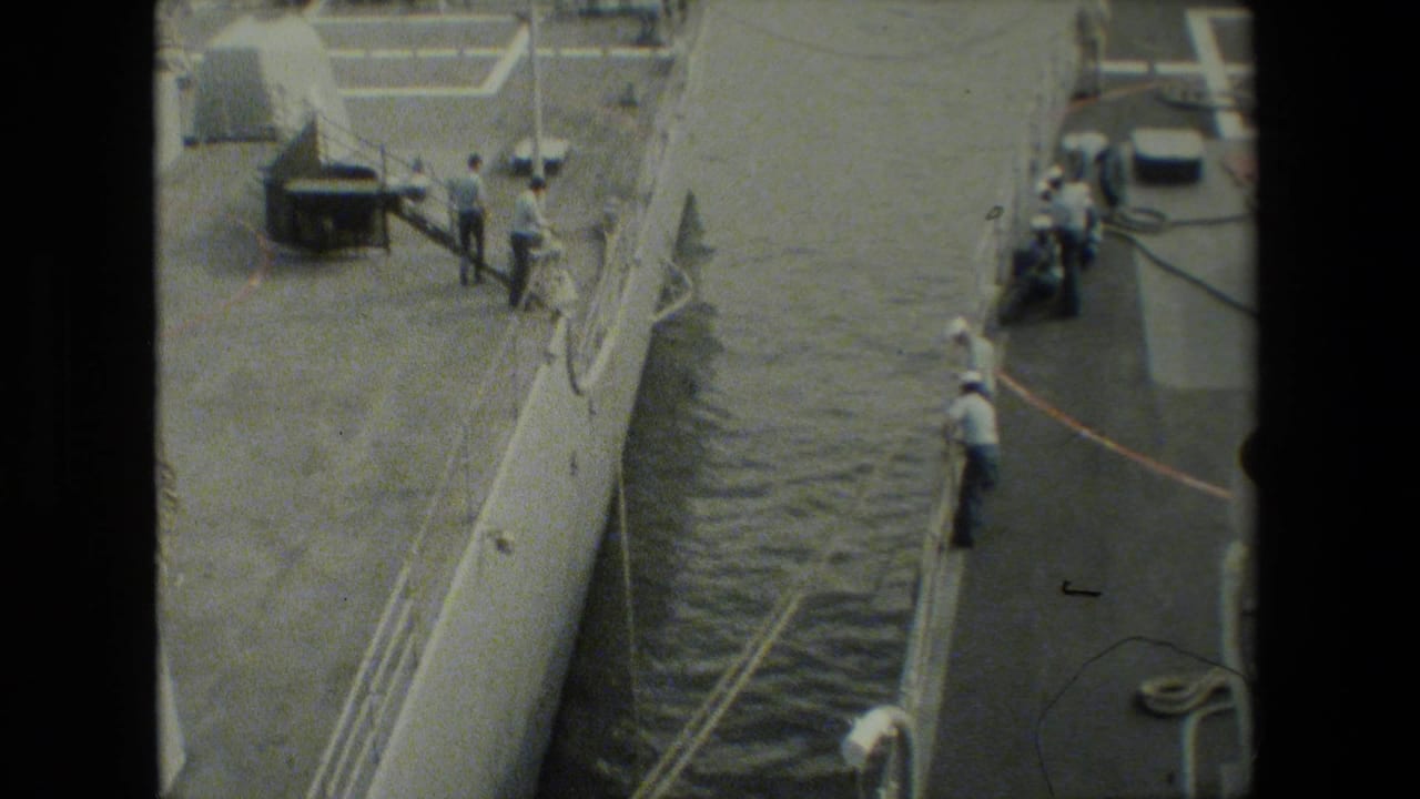 High-angle view of a moored military vessel, dock edge, and sailors in white uniforms appearing to handle lines or equipment beside the ship.