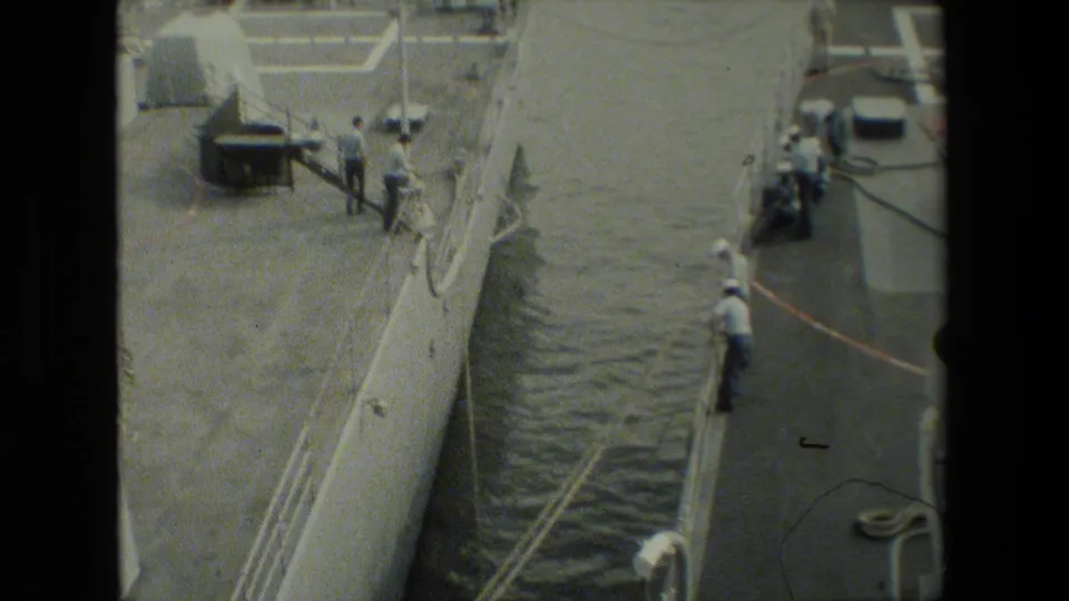 High-angle view of a moored military vessel, dock edge, and sailors in white uniforms appearing to handle lines or equipment beside the ship.