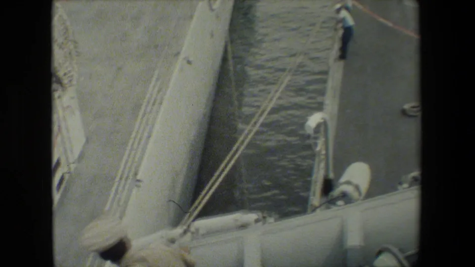Soft 8mm frame showing the side of a gray military ship, water between vessel and dock, and a crew member standing near the dock edge.