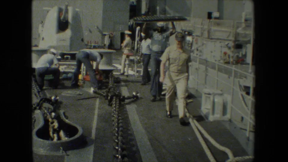 Deckside 8mm view of sailors and workers around chains, hoses, and equipment on or beside a military ship, with railings and vessel structure visible.