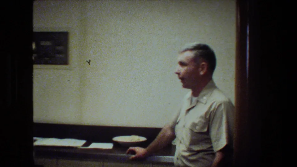 A uniformed man appears in profile inside a ship cabin or interior room, with a counter, plate, papers, and plain wall visible in soft 8mm footage.