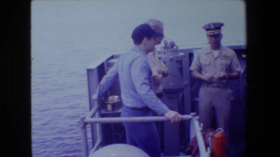 A sailor in a blue shirt and cap stands by the rail while a uniformed naval officer faces him on deck, with open water and deck equipment behind them.