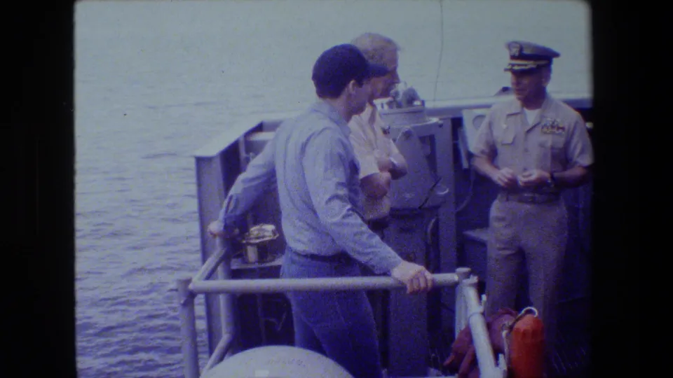 On a ship deck, a blue-shirted sailor leans near the railing while a uniformed officer stands opposite him, with water and deck fixtures visible in the background.