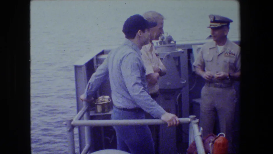 Adult men on a naval vessel deck stand near metal railings and ocean water, with one uniformed officer facing two men during a quiet exchange supported by the 1981 metadata.