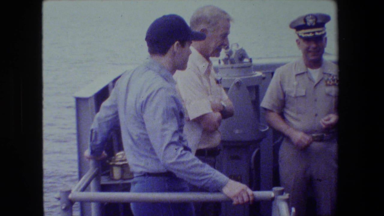 A man in a blue shirt and cap stands by a ship railing while a uniformed officer faces him on deck.