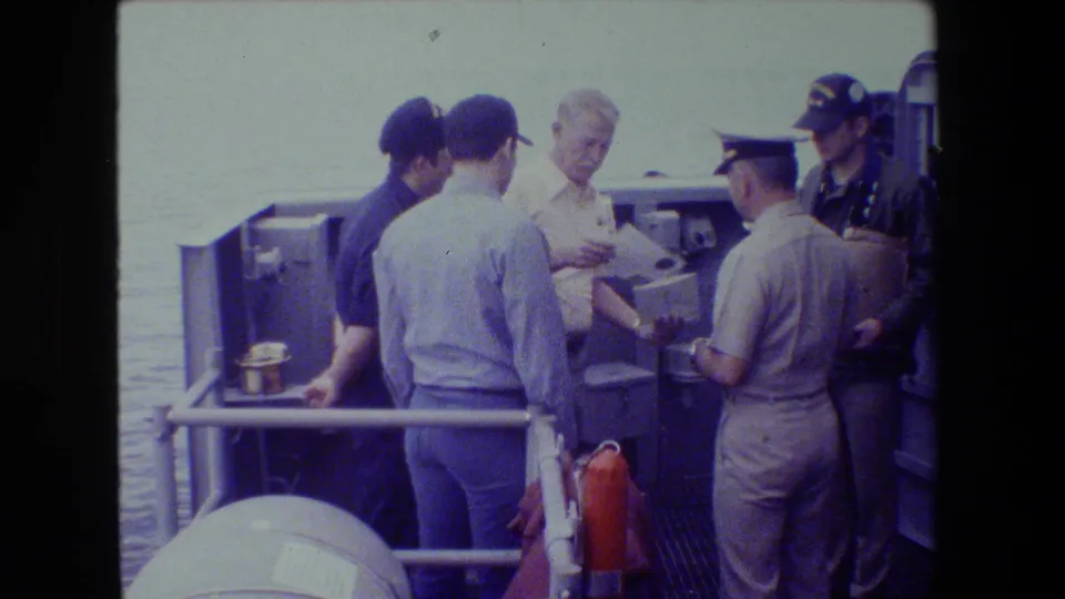 Several adult men cluster around a document or letter on a naval deck, including a uniformed man seen from behind and others in caps near the railing.