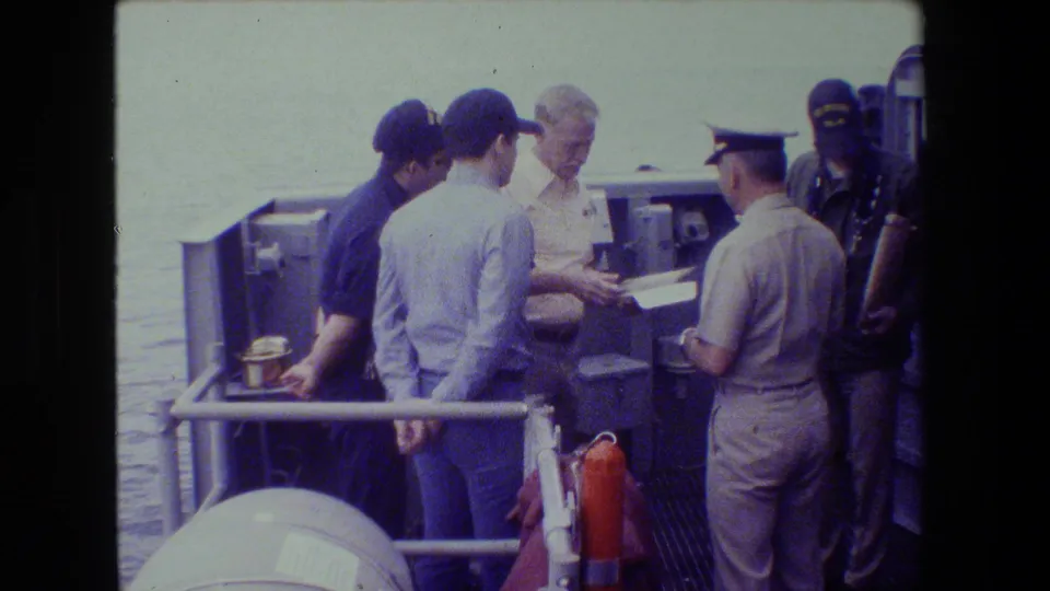 A man in a light shirt appears to hold or pass a document while uniformed and casually dressed men stand nearby on a naval vessel deck.