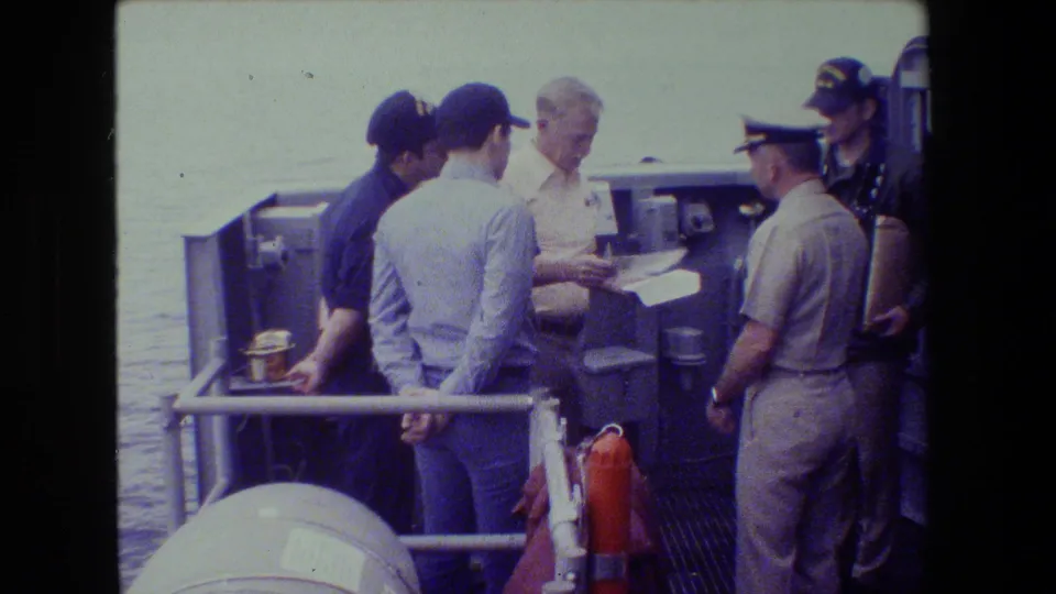 In 1981 archival 8mm footage, a uniformed naval officer appears to review a paper with crew members gathered on a ship deck at a naval setting supported by metadata.