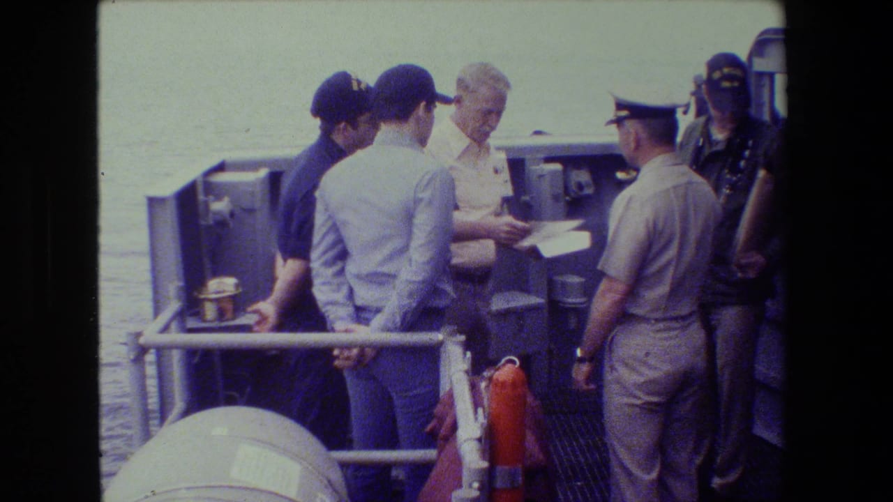 A naval officer in a light uniform stands with several men on a ship deck as they focus on a document, with water visible behind the railing.