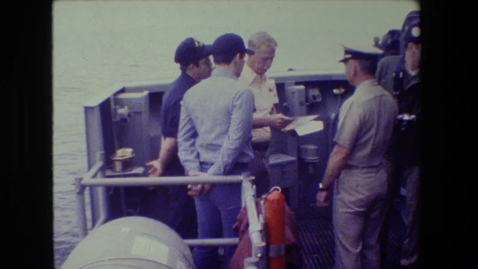 The frame shows an apparent shipboard briefing, with a light-uniformed officer and crew members gathered closely around paperwork on deck.
