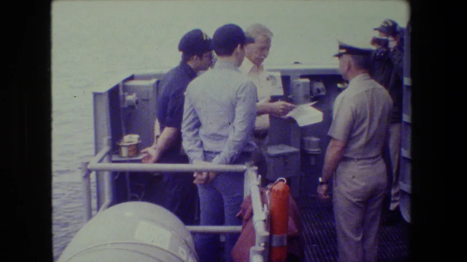 A naval officer appears to gesture over documents while crew members stand around him on a ship deck, captured with faded 8mm color and soft handheld framing.