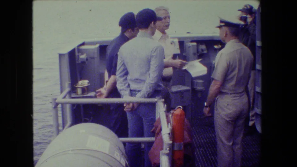 On a ship deck, a light-uniformed officer appears to present a document to men gathered near railings and deck equipment, consistent with the supplied naval ceremony metadata.