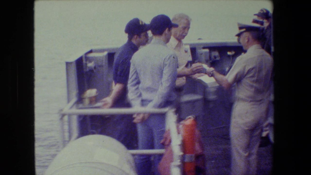 A uniformed officer holds papers while several men stand together on a deck by the water.